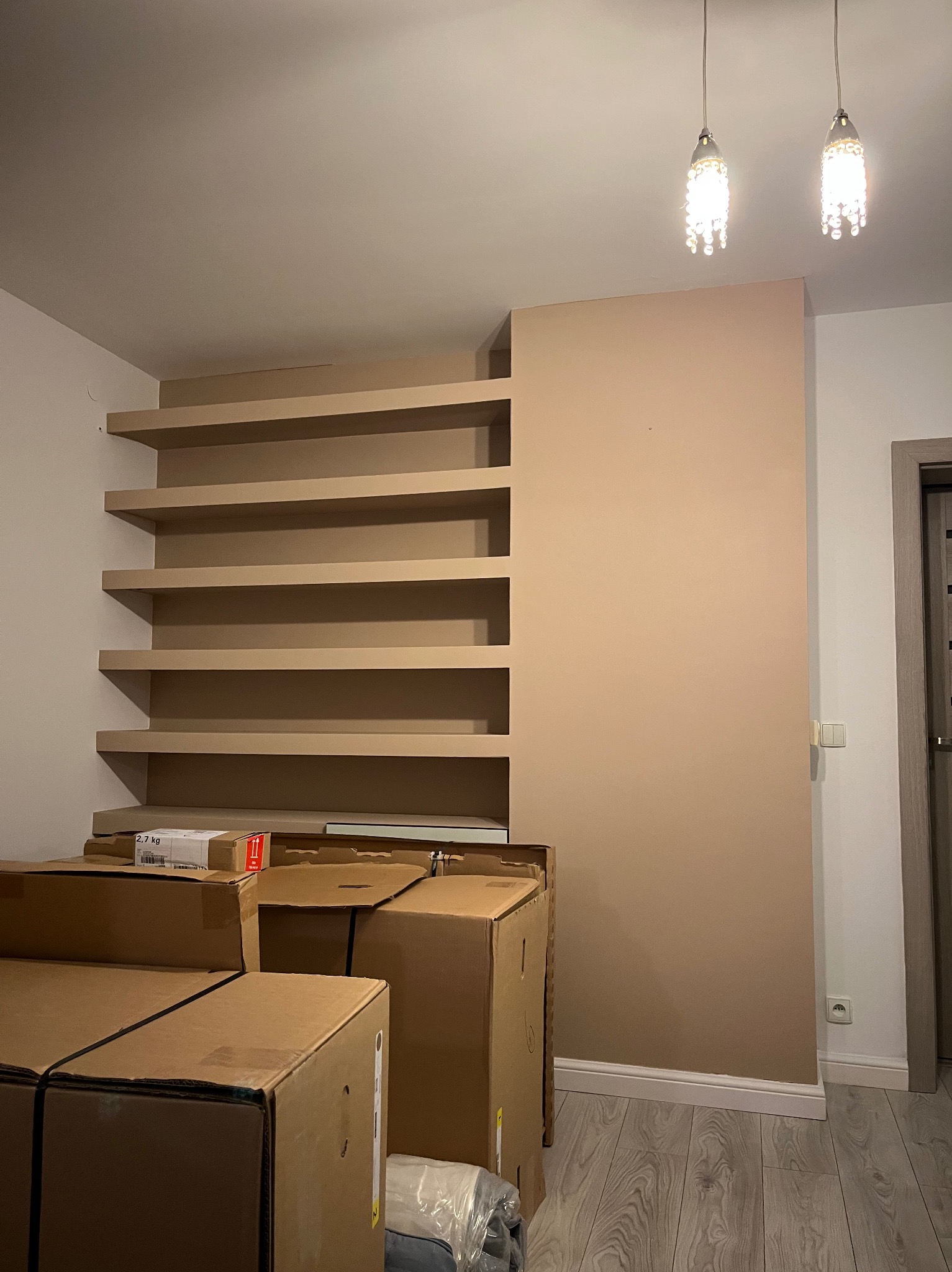 Pomalowana ściana z wbudowanymi półkami w kolorze beżowym, częściowo zasłonięta stertą kartonów na podłodze z panelami, dwie wiszące lampy w tle.