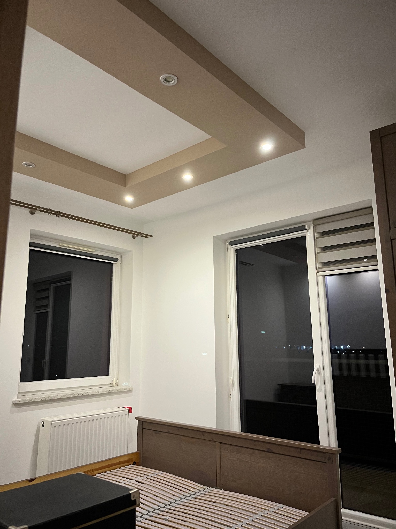 Sypialnia po remoncie: widok na sufit z dekoracyjnym podwieszanym elementem z oświetleniem punktowym, łóżko z drewnianym wezgłowiem, grzejnik pod oknem, rolety w oknach.