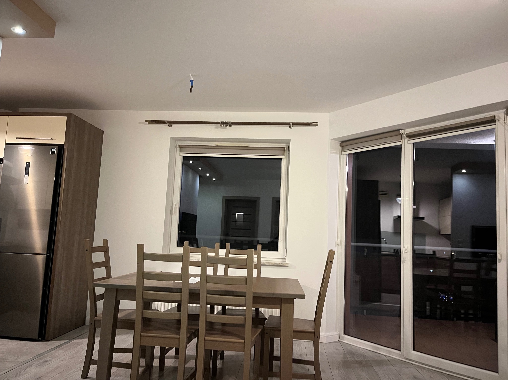 Jasne wnętrze jadalni z drewnianym stołem i krzesłami, widok na okno z roletami i przesuwne drzwi balkonowe, na ścianie widoczny ślad po taśmie malarskiej.