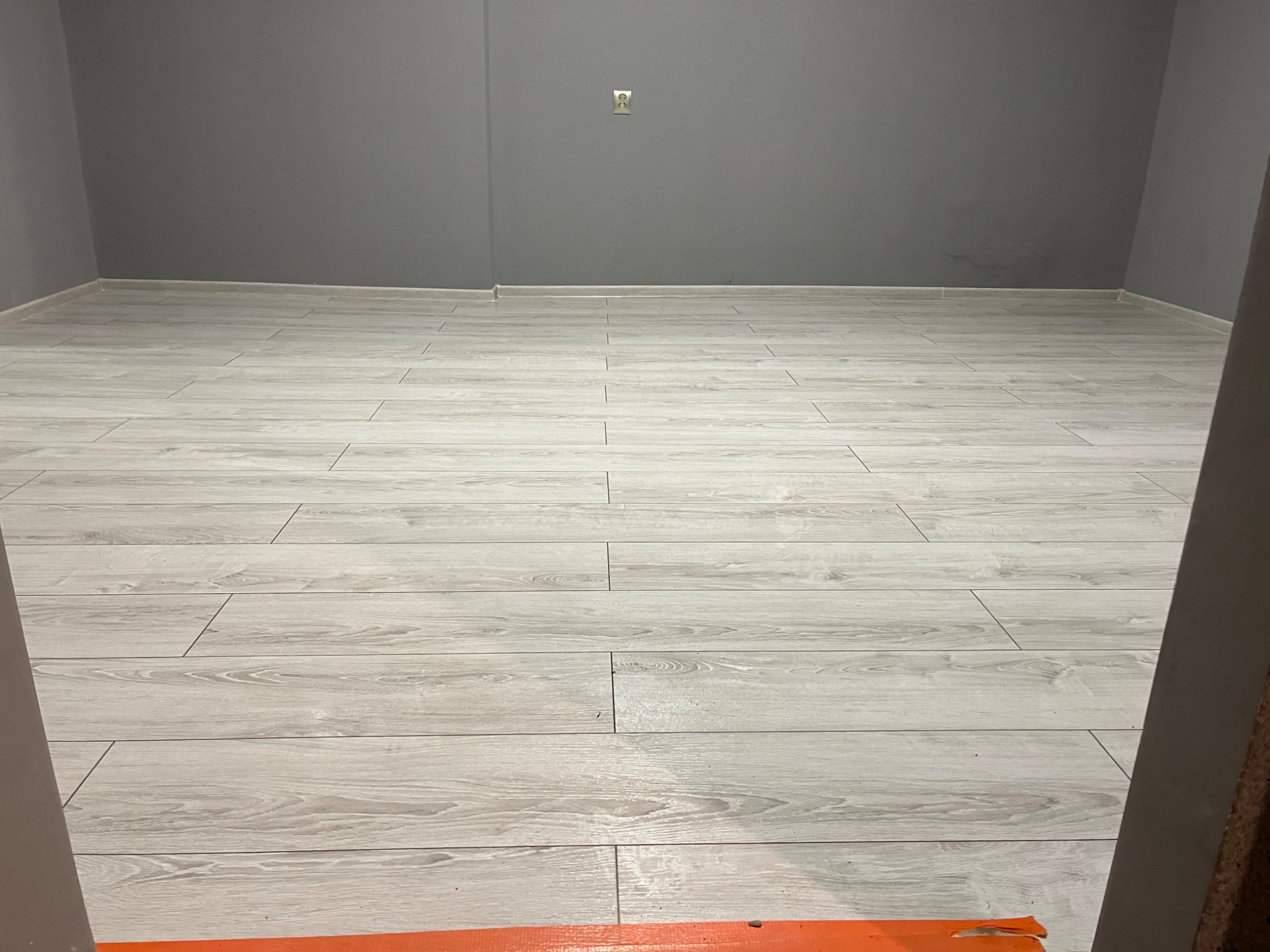 Wykończone wnętrze z jasnymi panelami podłogowymi imitującymi drewno, szare ściany i widoczne gniazdko elektryczne, fragment ościeżnicy w prawym dolnym rogu.