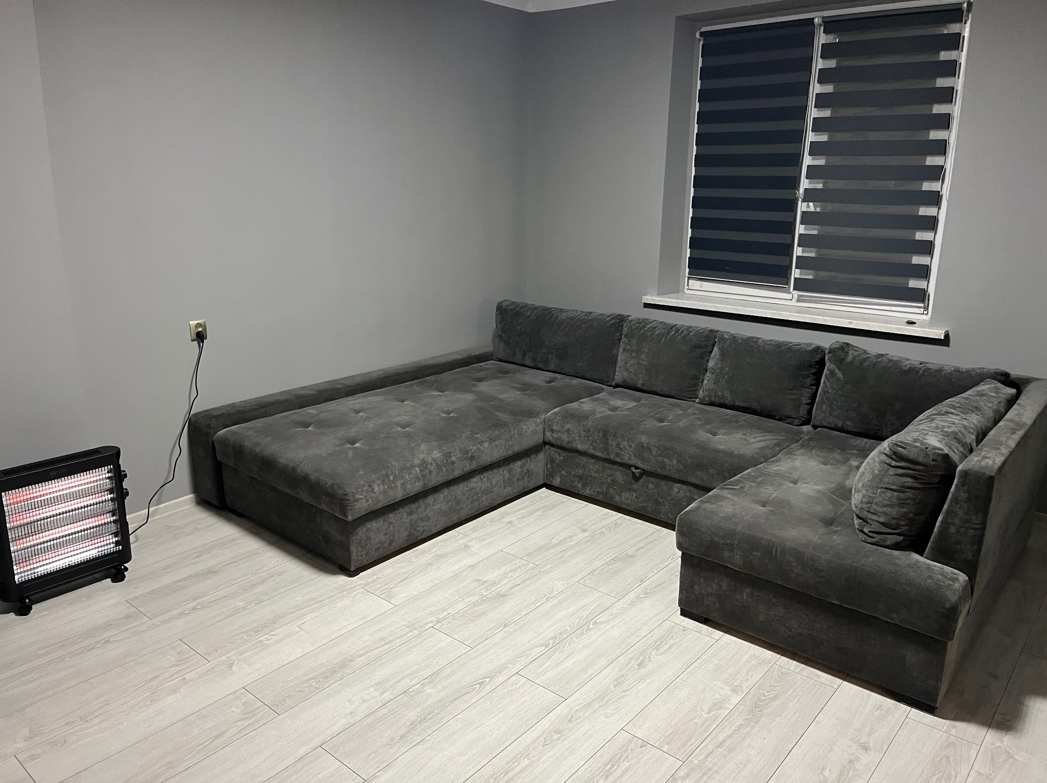 Szary narożnik z funkcją spania w pokoju o szarych ścianach i jasnej podłodze, widoczne rolety w oknie i grzejnik elektryczny w rogu.