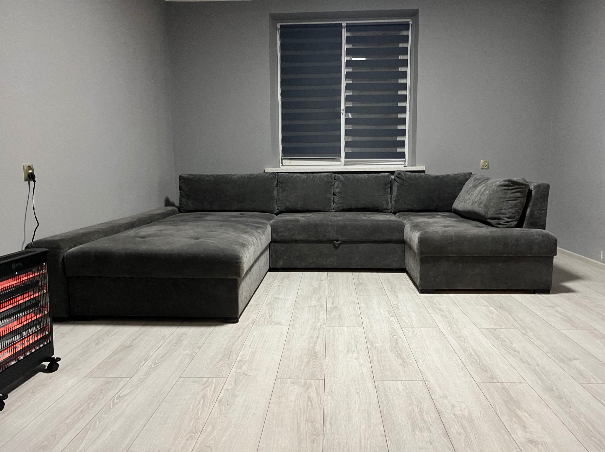 Szary, welurowy narożnik z funkcją spania na jasnej podłodze z paneli, ogrzewany przez elektryczny grzejnik halogenowy, szare ściany, okno z żaluzjami dzień-noc.