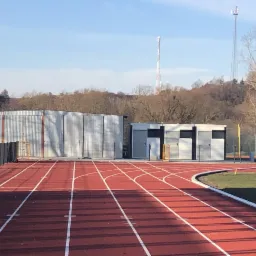 Widok z poziomu bieżni na halę sportową z blachy falistej oraz budynek zaplecza sanitarnego na tle zalesionego wzgórza i wież telekomunikacyjnych.