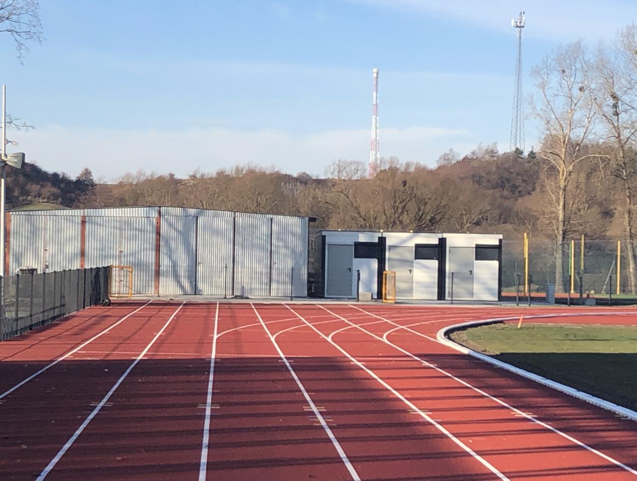 Widok z poziomu bieżni na halę sportową z blachy falistej oraz budynek zaplecza sanitarnego na tle zalesionego wzgórza i wież telekomunikacyjnych.