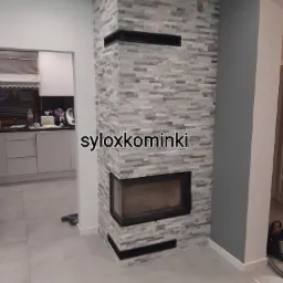 Narożny kominek obłożony szarą, łupkową okładziną, z czarnymi elementami wykończeniowymi, widoczny w tle fragment białej kuchni i szarej ściany.