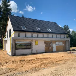 Surowy stan domu z białego betonu komórkowego z czarnym dachem i trzema oknami dachowymi, widoczne żółte rury wentylacyjne, teren wokół budynku pokryty piaskiem, w tle zieleń drzew.