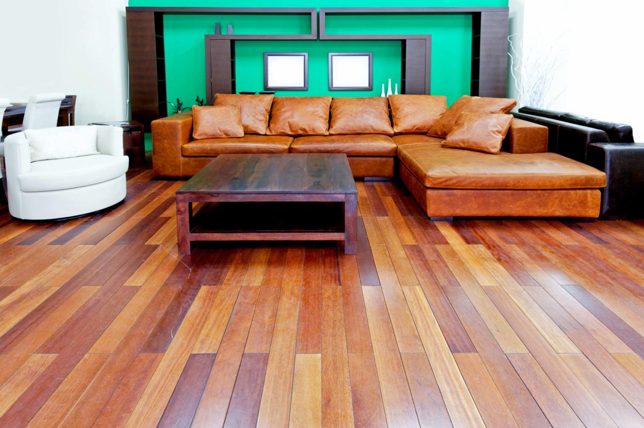 Brązowy narożnik skórzany z poduchami w salonie z drewnianą podłogą i stolikiem kawowym, na tle zielonej ściany z półkami.