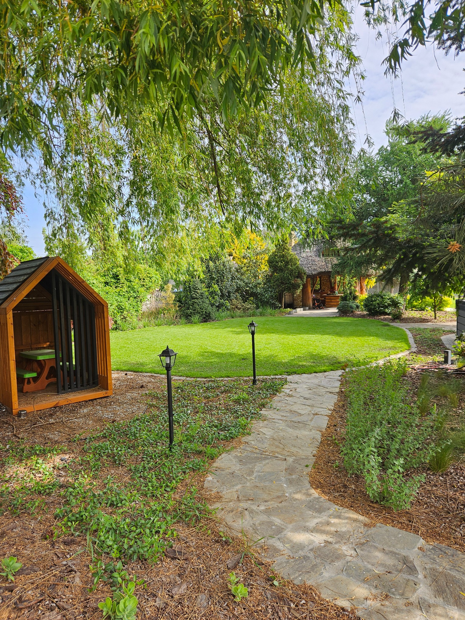 Urokliwy ogród z kamienną ścieżką, altaną krytą strzechą i małą drewnianą budką z zielonym stołem, otoczony zielenią i drzewami.