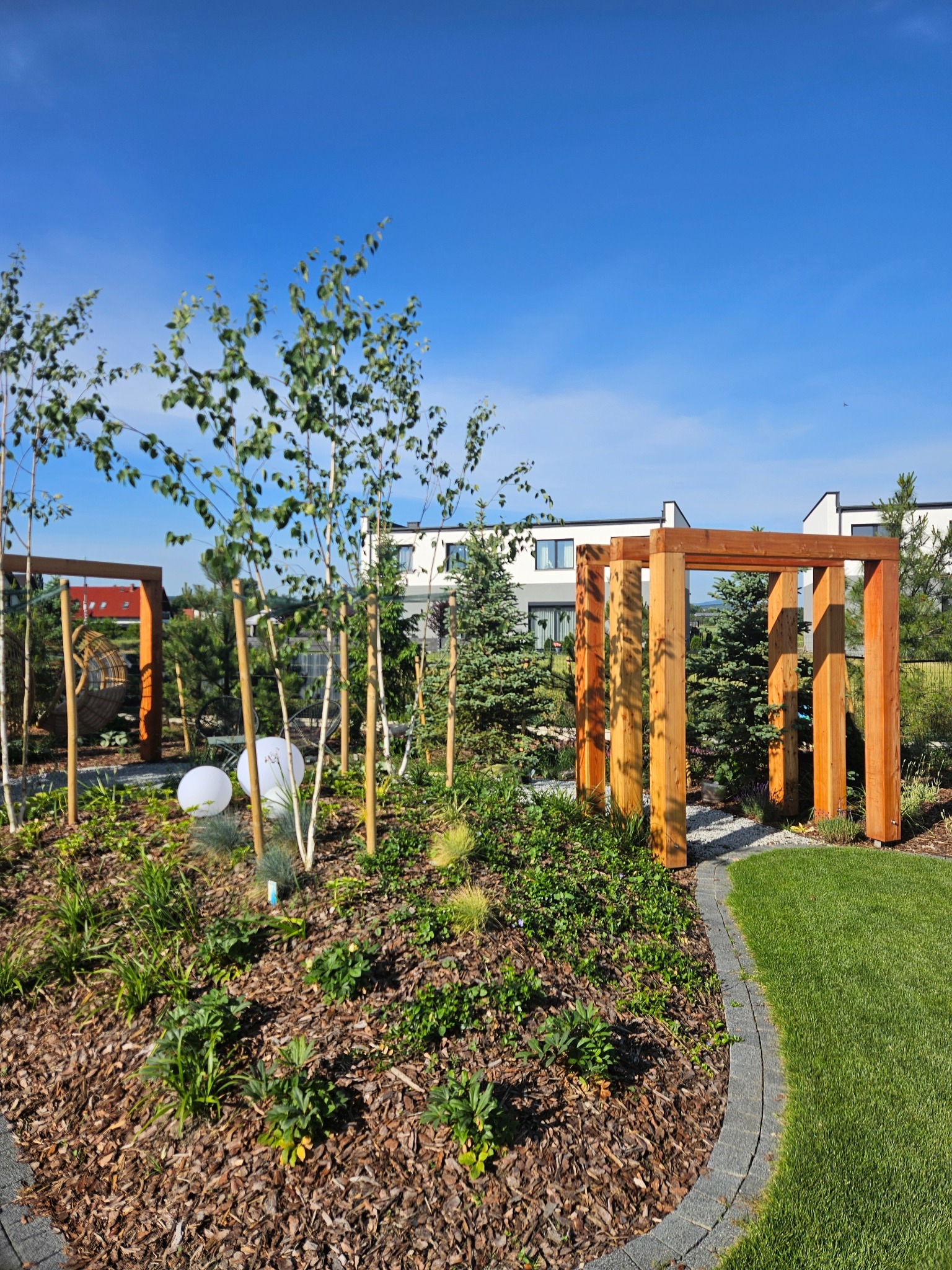 Nowoczesny ogród z drewnianą pergolą, korą, trawą i młodymi drzewami na tle białego domu w słoneczny dzień. Elementy oświetlenia ogrodowego.