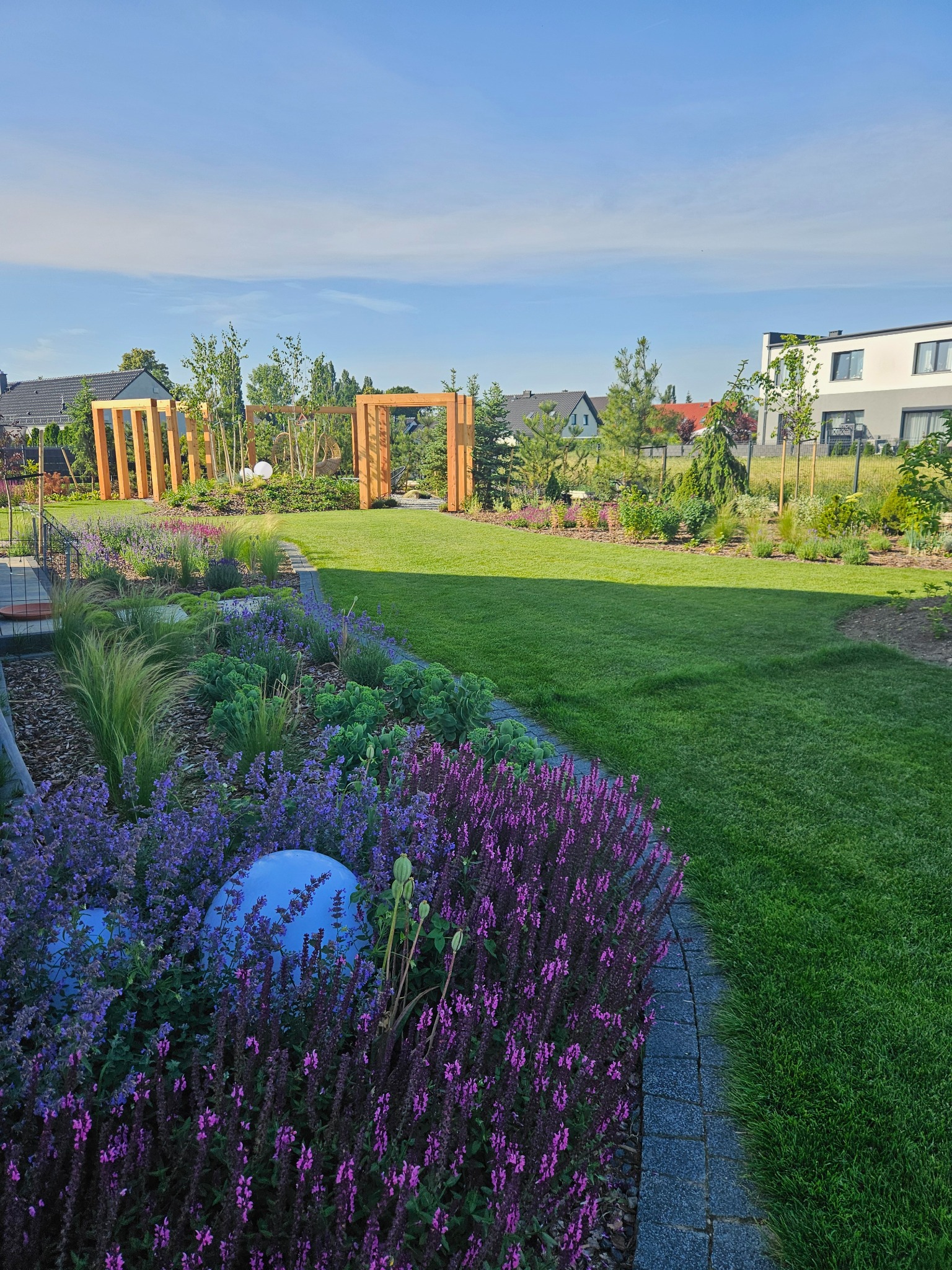 Urokliwy ogród z drewnianymi pergolami, kwitnącymi rabatami w odcieniach fioletu i zieleni, oraz idealnie przystrzyżonym trawnikiem. Dekoracyjne kule dodają nowoczesnego akcentu.