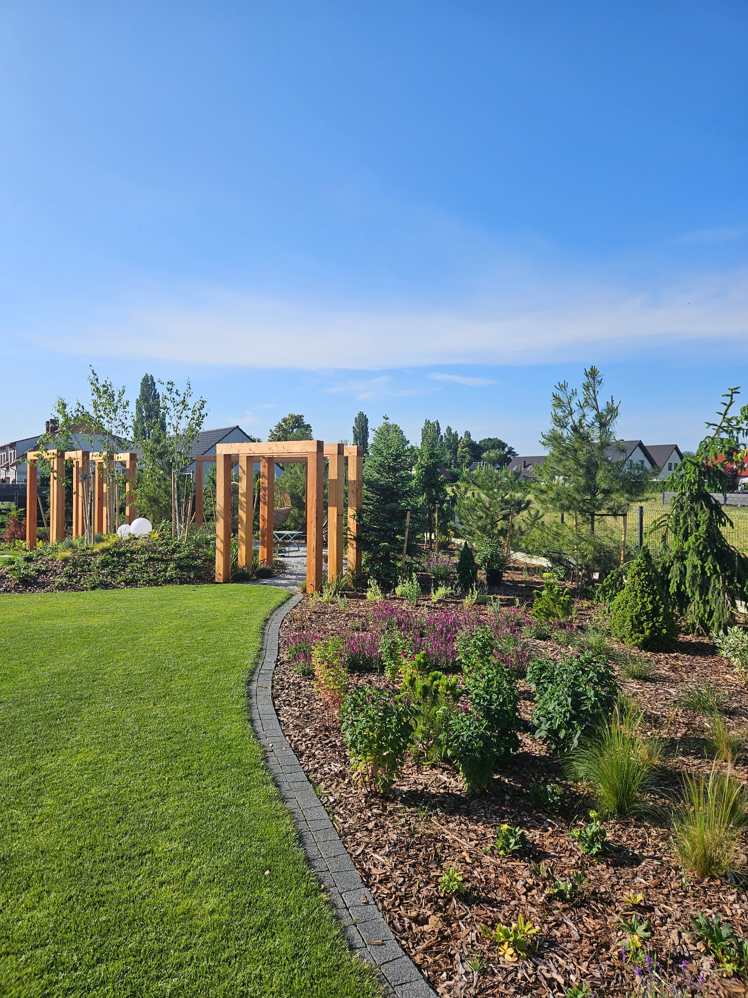 Zadbany ogród z drewnianą pergolą, rabatami kwiatowymi i równo przystrzyżonym trawnikiem. Widoczne drzewa iglaste i liściaste, kostka brukowa oddzielająca trawnik od rabat.