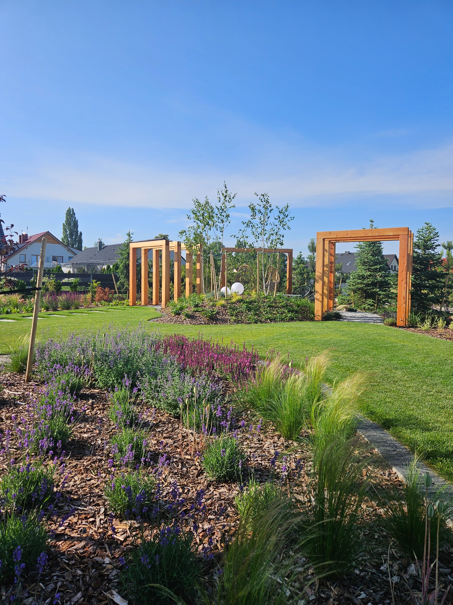 Starannie zaprojektowany ogród z drewnianymi konstrukcjami, rabatami kwiatowymi i trawnikiem, w słoneczny dzień. Widoczne elementy architektury ogrodowej.