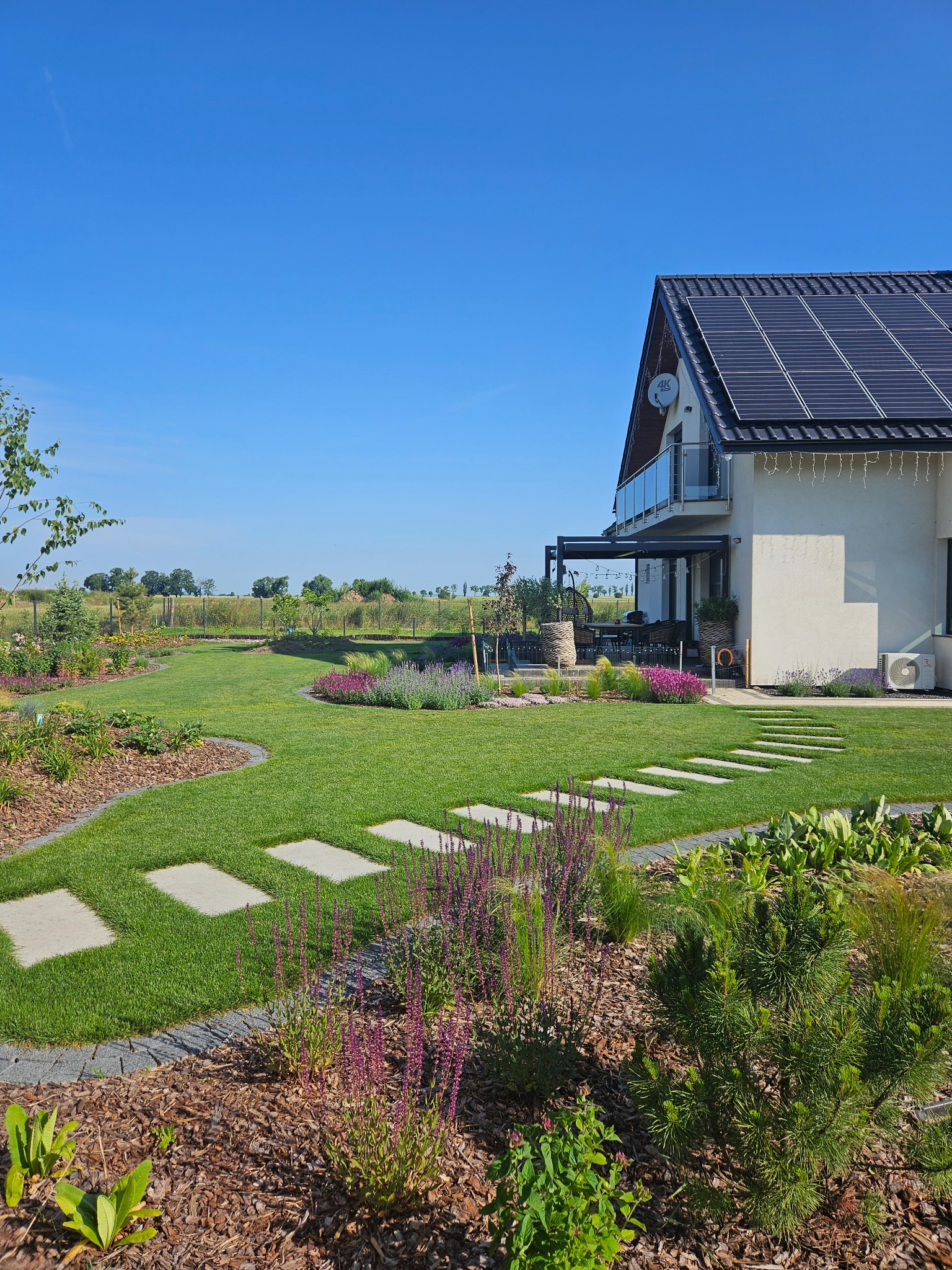 Nowoczesny ogród z trawnikiem, ścieżką z płyt, rabatami kwiatowymi i drzewami, na tle domu z panelami słonecznymi w słoneczny dzień. Widok na zadbany teren.