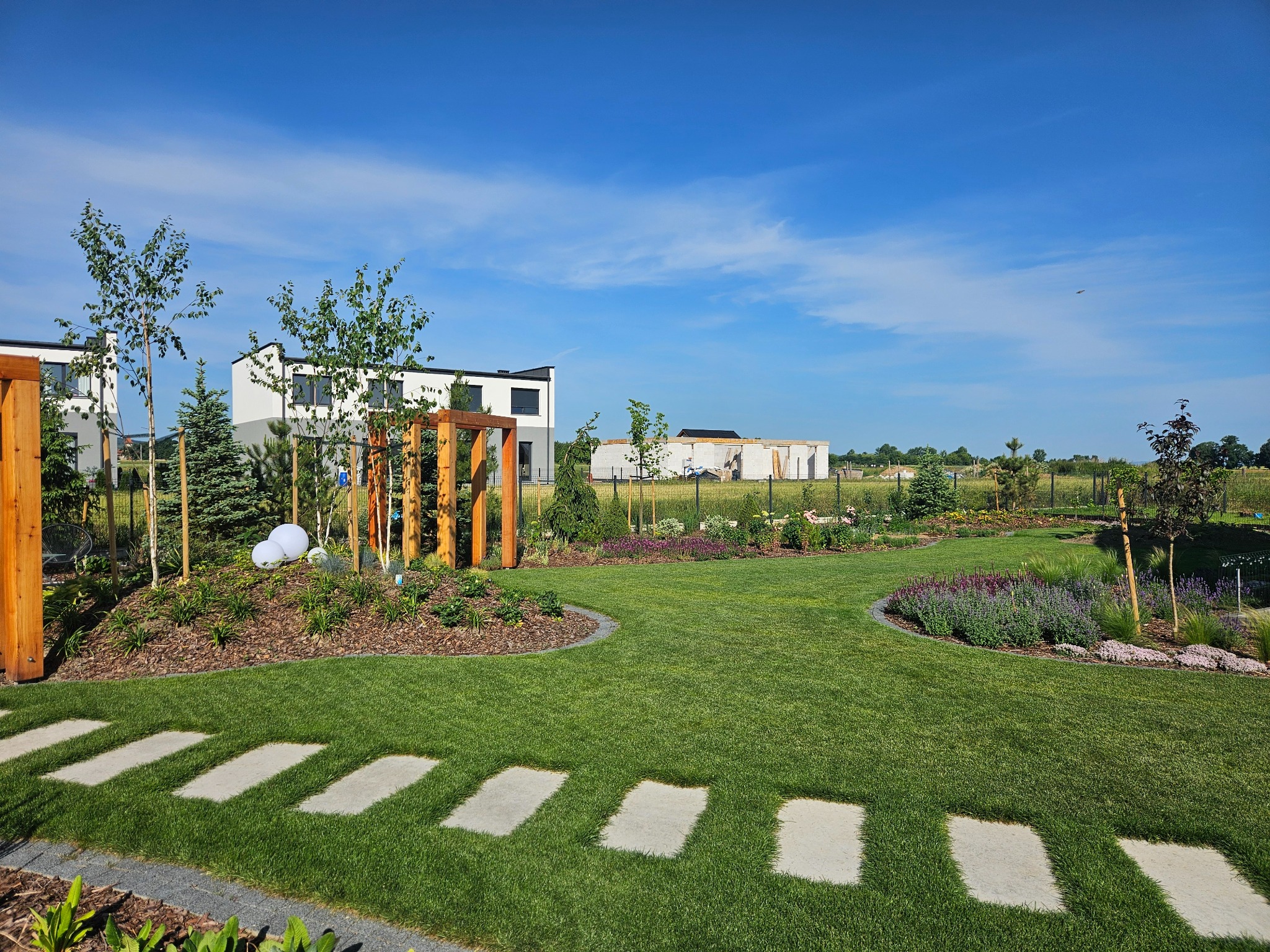 Zadbany ogród z równo przystrzyżonym trawnikiem, ścieżką z płyt, rabatami kwiatowymi i ozdobnymi drzewami na tle nowoczesnych domów i błękitnego nieba.