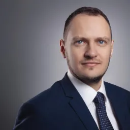 Portret mężczyzny w ciemnym garniturze i krawacie w kropki, z chusteczką w kieszonce, na szarym tle.