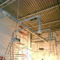 Skomplikowany system wentylacyjny z metalowych rur zamontowany pod sufitem hali, widoczne rusztowanie i drabina.