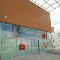 Elewacja biurowca z drewnianą okładziną na górnej części, przeszklona dolna część z widocznymi elementami konstrukcyjnymi wewnątrz, plakaty na szybach.