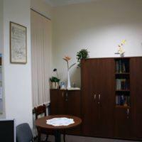 Neutralne wnętrze pokoju z drewnianymi meblami, okrągłym stolikiem z serwetą i dekoracjami roślinnymi na szafkach. Na ścianie wisi oprawiony obrazek.