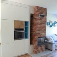Nowoczesna kuchnia z białymi, matowymi szafkami w zabudowie, z wbudowanym piekarnikiem i elementami z drewna oraz błękitnymi półkami, sąsiadująca z ceglanym kominkiem i fragmentem salonu z szarą sofą.