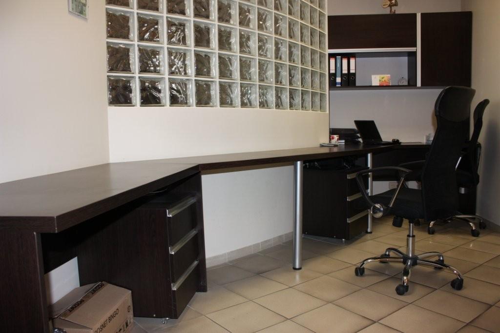 Nowoczesne meble biurowe w Szczecinie: Długi, narożny stół z ciemnego drewna z metalowymi nogami i szafkami z szufladami, uzupełniony o ergonomiczne krzesła biurowe na kółkach, na tle ściany...