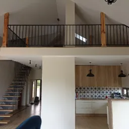 Przestronne wnętrze domu z antresolą i drewnianymi schodami z metalową balustradą, widok na kuchnię z drewnianymi frontami i niebiesko-białymi płytkami oraz wiszącymi lampami.