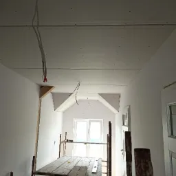 Montaż płyt kartonowo-gipsowych na suficie poddasza, widoczne kable elektryczne, rusztowanie robocze i okno w tle.