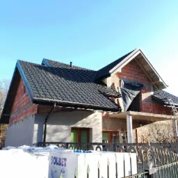 Wznoszony dom z częściowo ukończonym dachem z czarnej dach&oacute;wki, widoczne bloczki Solbet przed posesją i surowy stan elewacji.