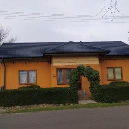 Usługi Budowlane Bartosz Ziółczyk - Pomarańczowy dom jednorodzinny z nowym, ciemnoszarym dachem z dachówki, z numerem 24 i wejściem ozdobionym łukiem z zielonych pnączy, widoczne fragmenty elewacji w trakcie ocieplania.
