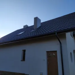 Nowoczesny dom jednorodzinny z ciemną, metalową dachówką, widoczne kominy, okno dachowe i orynnowanie, w tle błękitne niebo.