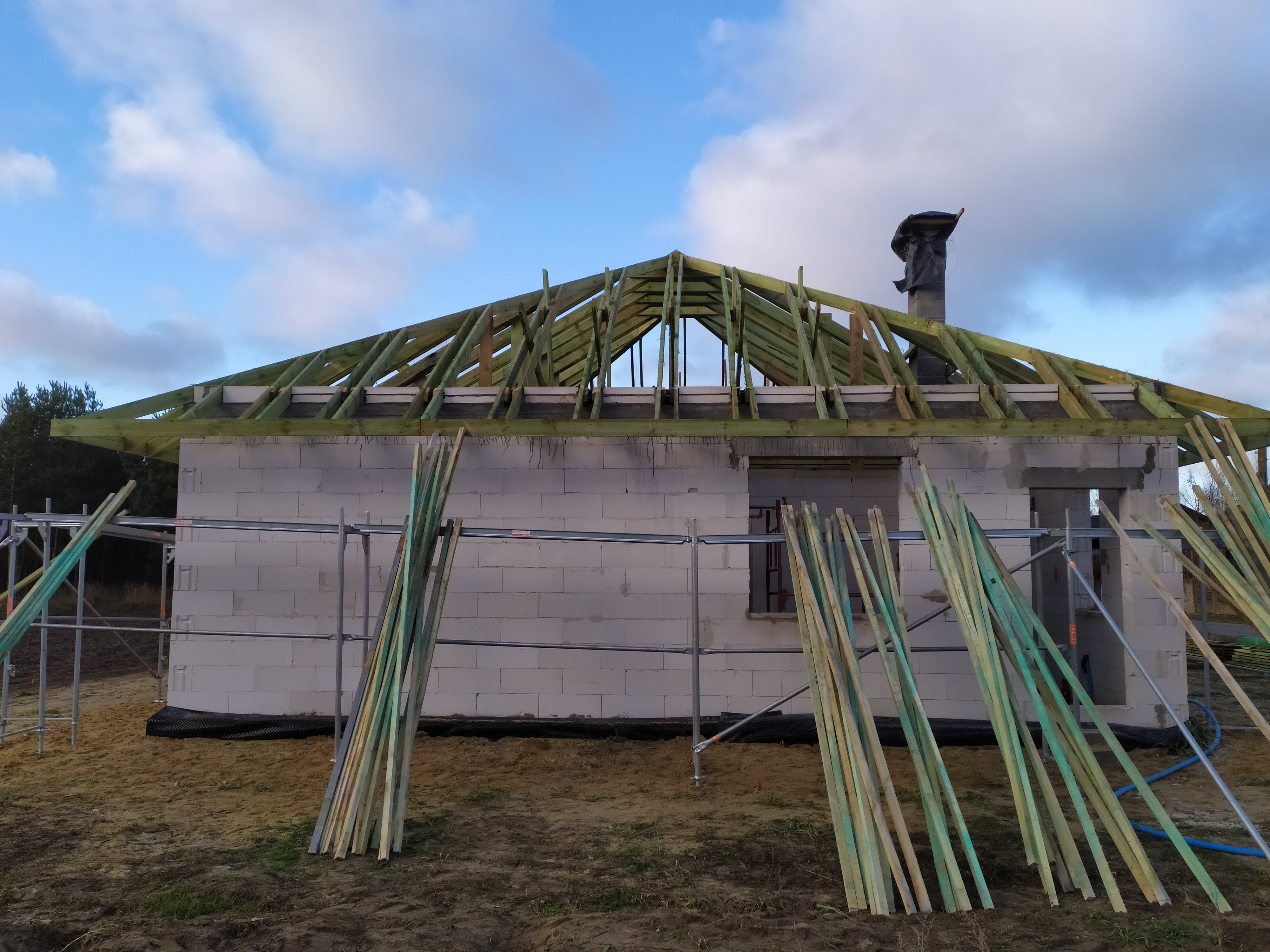 Budowa domu z białych bloczków: widoczna konstrukcja drewniana dachu, rusztowania i stos desek obok budynku.