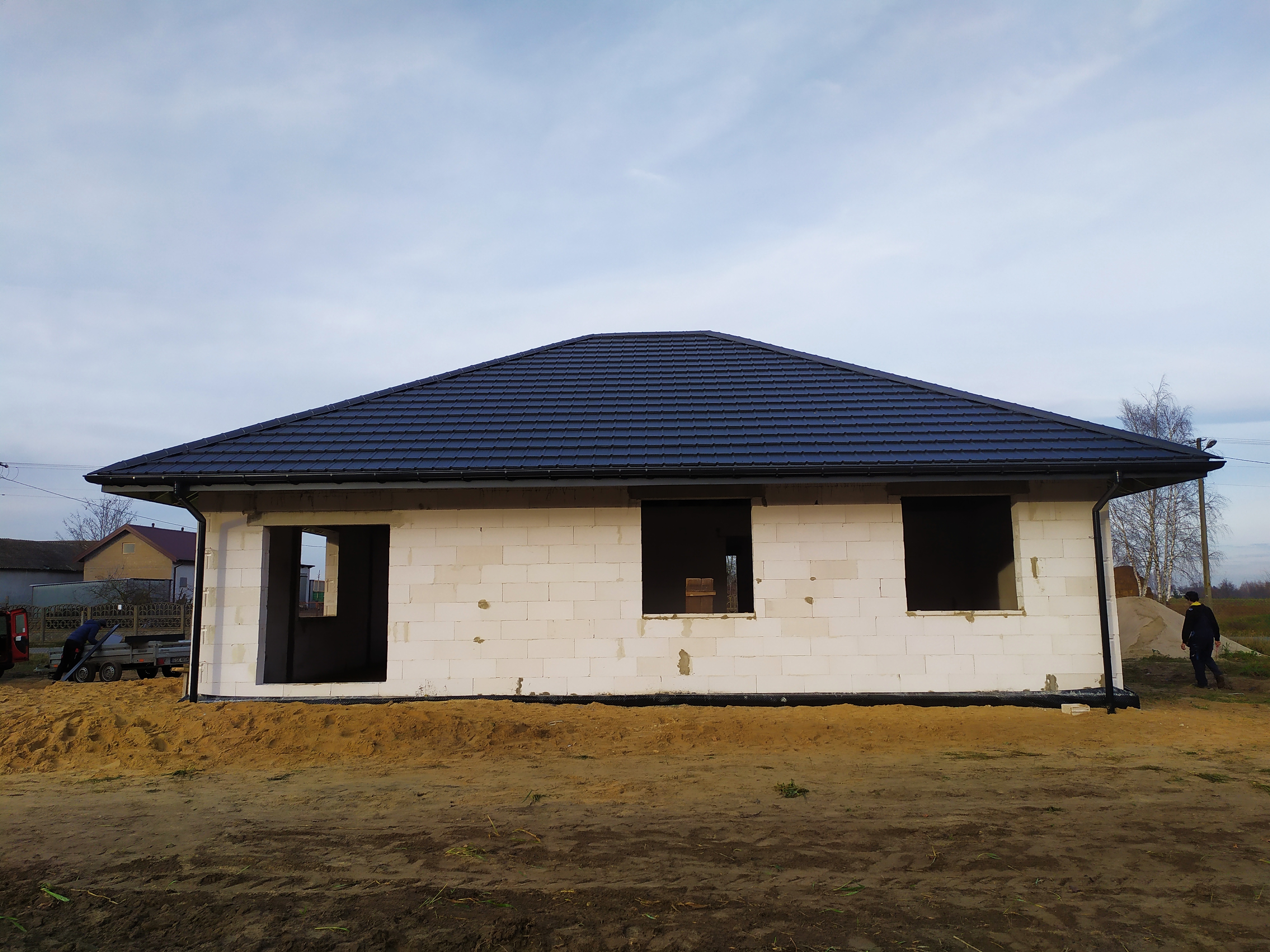 Surowy stan domu jednorodzinnego z białego bloczka, z zamontowanym, ciemnym dachem z dachówki ceramicznej, przygotowany do dalszych prac wykończeniowych, widoczne otwory okienne bez stolarki.