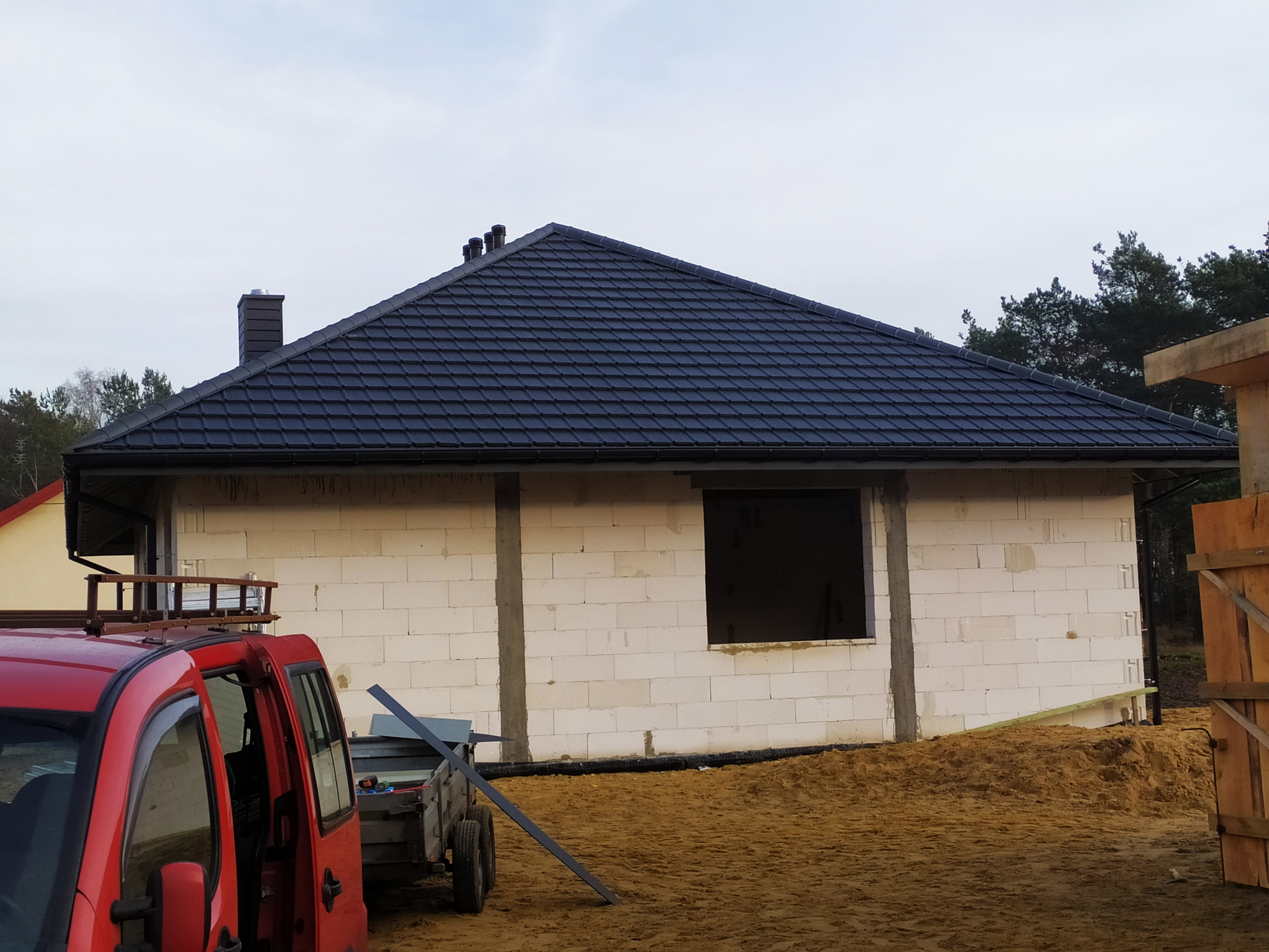 Niedokończony dom z białych bloczków z czarnym dachem z dachówki, widoczny samochód dostawczy z drabinką na dachu i przyczepką z elementami blacharskimi.