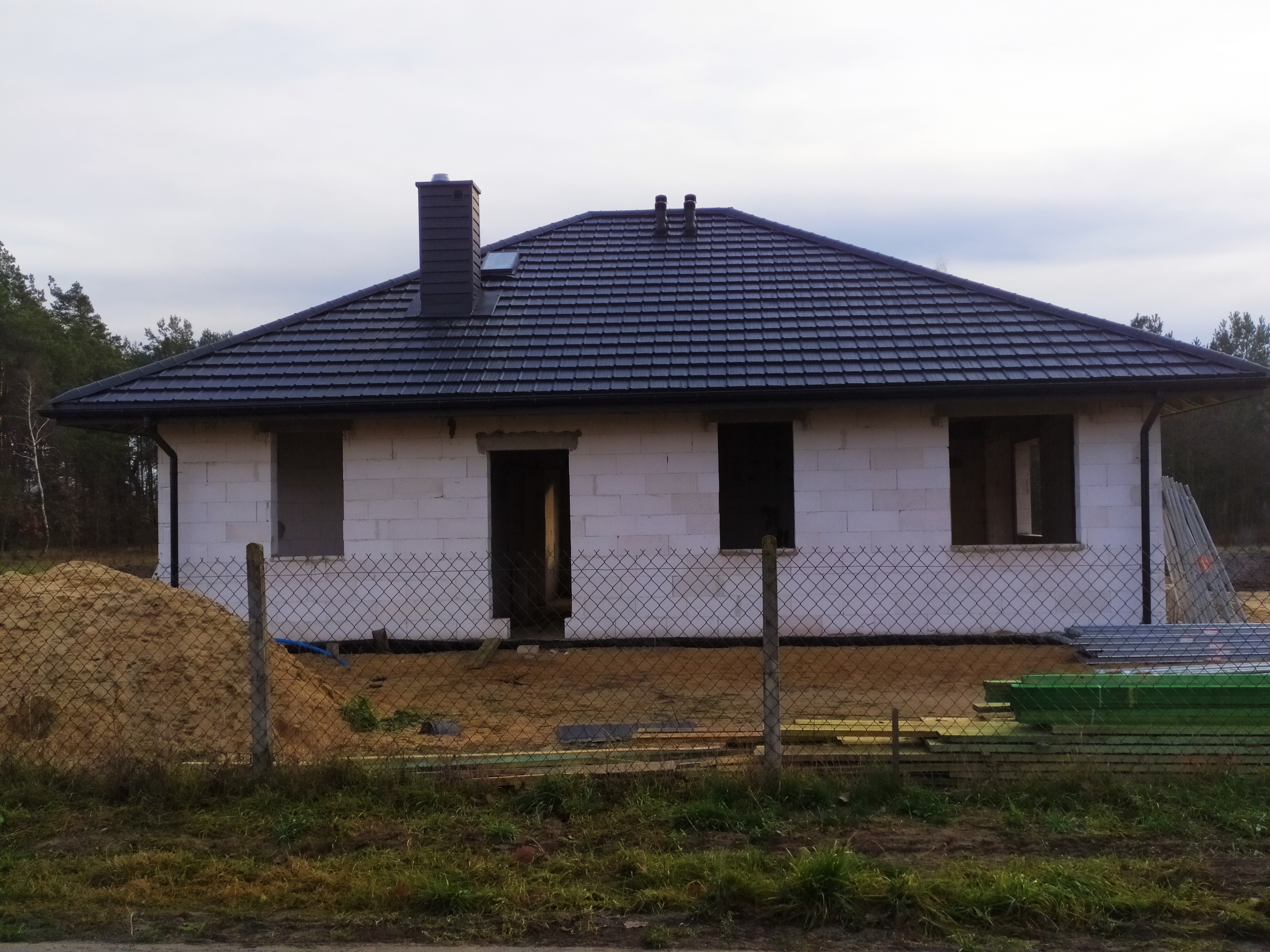 Niedokończony dom z białego pustaka z nowym, ciemnym dachem ceramicznym, widoczne kominy, brak okien i drzwi, materiały budowlane składowane na działce, ogrodzenie z siatki.