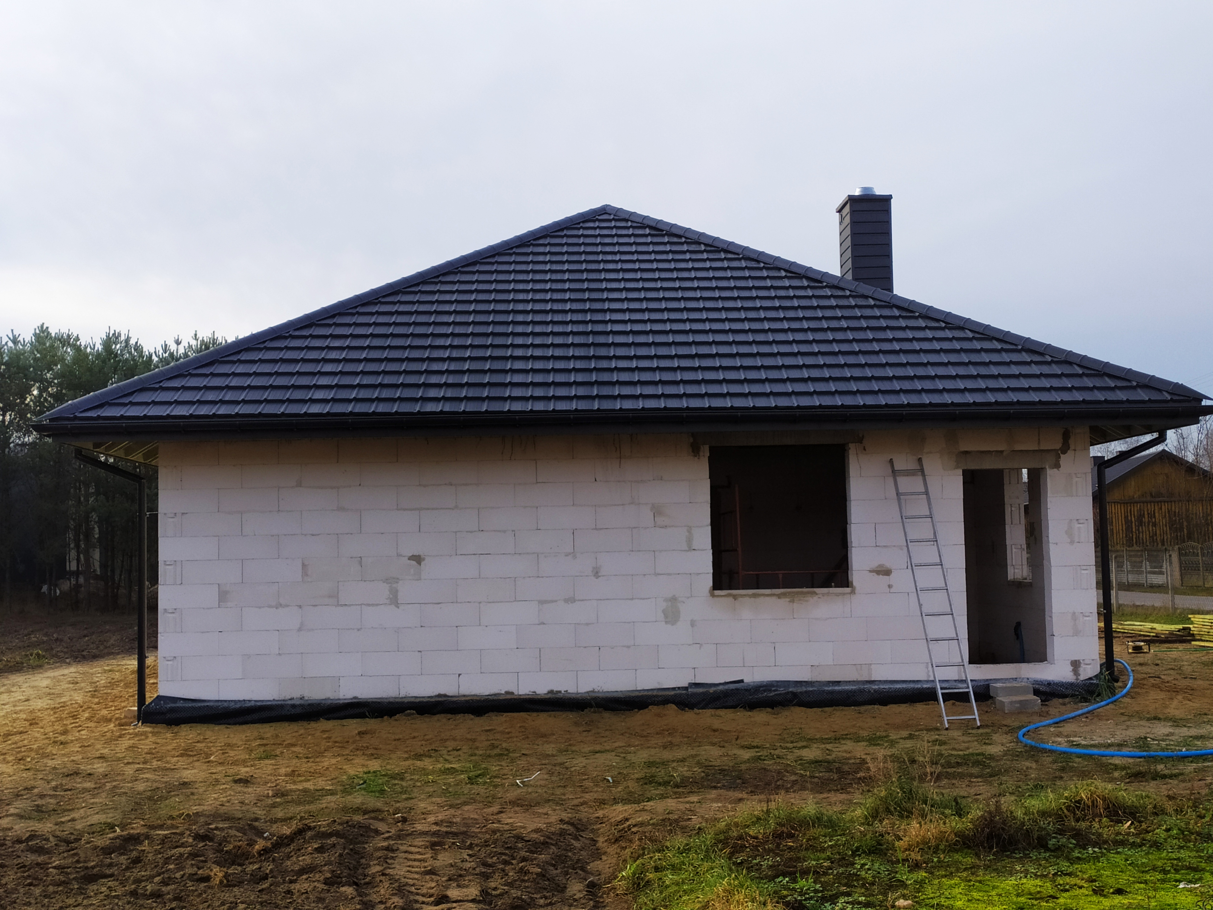 Surowy stan domu z białych bloczków z czarnym dachem z dachówki, widoczny komin, drabina oparta o ścianę, w tle drzewa.