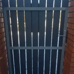 Ciemnoszara furtka z pionowych desek w metalowej ramie, zamontowana między ceglanymi słupkami, widoczny fragment chodnika z kostki brukowej.