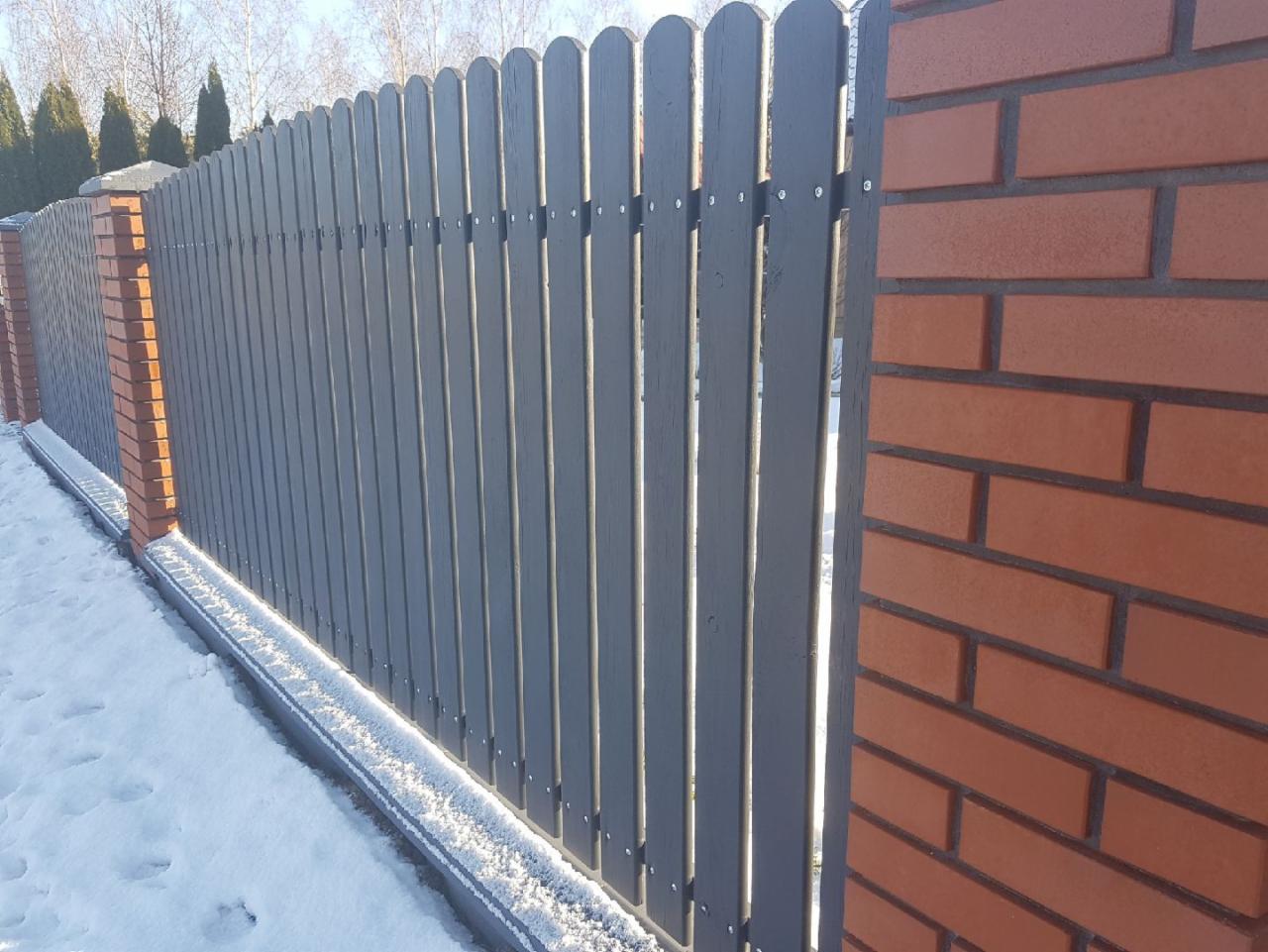 Szare drewniane ogrodzenie sztachetowe z ceglanymi słupkami w zimowej scenerii, lekko ośnieżone.