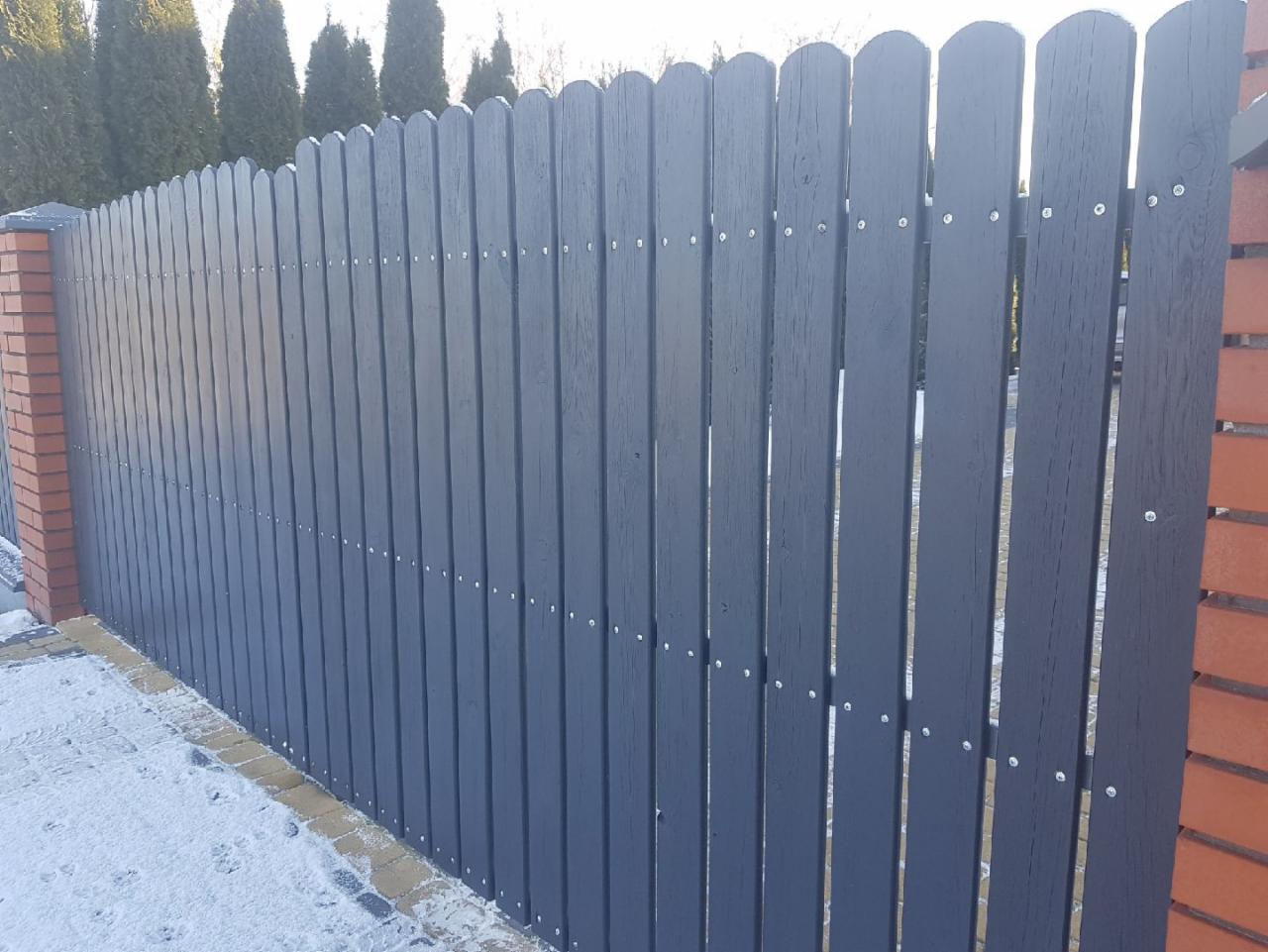 Grafitowe ogrodzenie z pionowych desek o zaokrąglonym zakończeniu, mocowane na śruby, z ceglanymi słupkami na tle zimowego krajobrazu.