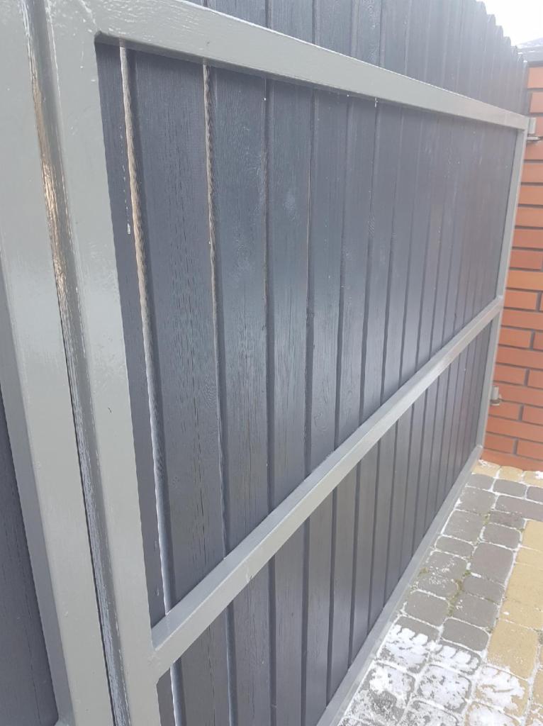 Fragment grafitowego ogrodzenia z pionowych desek, z szarą metalową ramą, widziany z boku, z fragmentem muru z cegły w tle.
