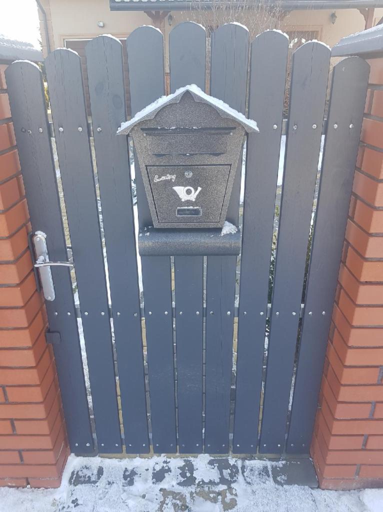 Ciemnoszara furtka z drewnianych desek z zamontowaną metalową skrzynką na listy, ośnieżona, między dwoma słupkami z czerwonej cegły.
