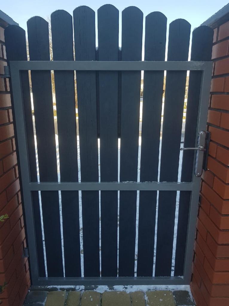 Ciemnoszara furtka z pionowych desek w metalowej ramie, zamontowana między ceglanymi słupkami, widoczny fragment chodnika z kostki brukowej.