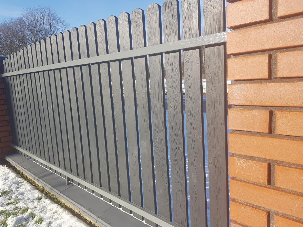 Szare pionowe deski ogrodzeniowe z metalowym poprzecznym wspornikiem, połączone z ceglanym słupkiem, widoczne resztki śniegu.