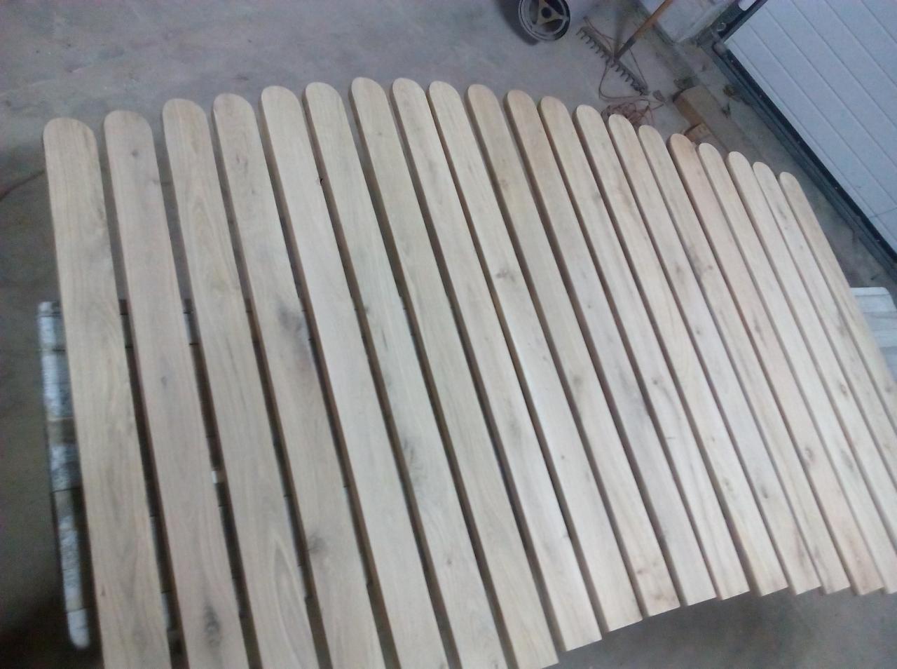 Surowe, drewniane sztachety z zaokrąglonymi końcami, ułożone równolegle na szarym podłożu w warsztacie, przed malowaniem lub montażem.