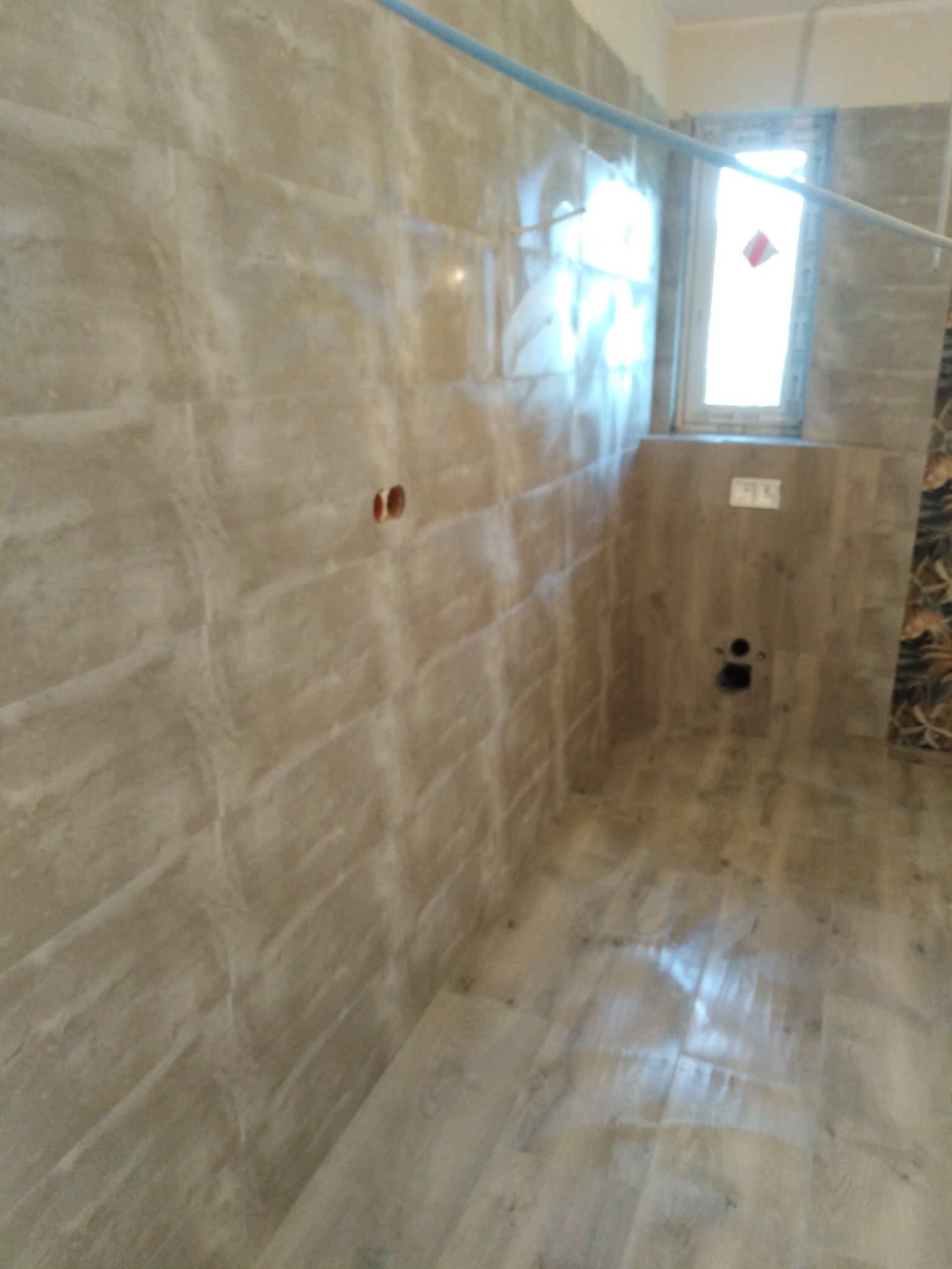 Wykończona łazienka z beżowymi płytkami imitującymi kamień na ścianach i podłodze, widoczne gniazdka elektryczne i rura na suficie.