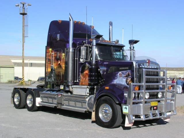 Fioletowy ciągnik siodłowy Kenworth z chromowanymi elementami i grafiką fantasy na kabinie, zaparkowany na placu.