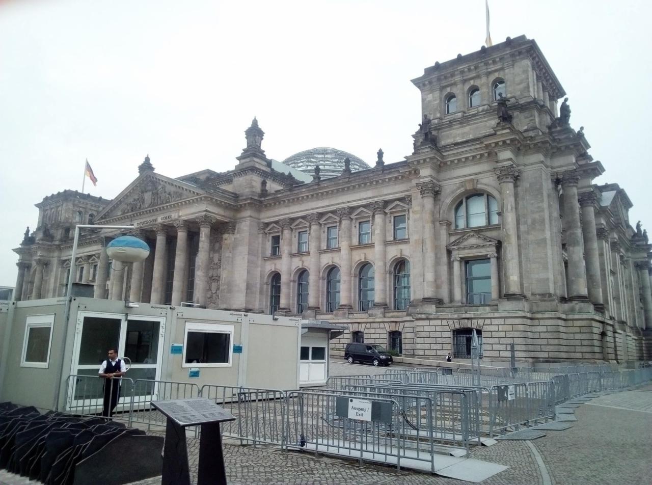 Fasada budynku Reichstagu w Berlinie z widocznymi kolumnami, kopułą i flagą, otoczona tymczasowymi kontenerami i barierkami z napisem 'Ausgang / Exit'.