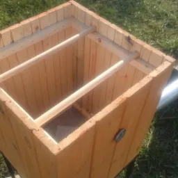 Drewniana skrzynia z rurą odprowadzającą, ustawiona na trawie, widok z góry, jasne drewno, widoczne elementy konstrukcyjne wewnątrz.