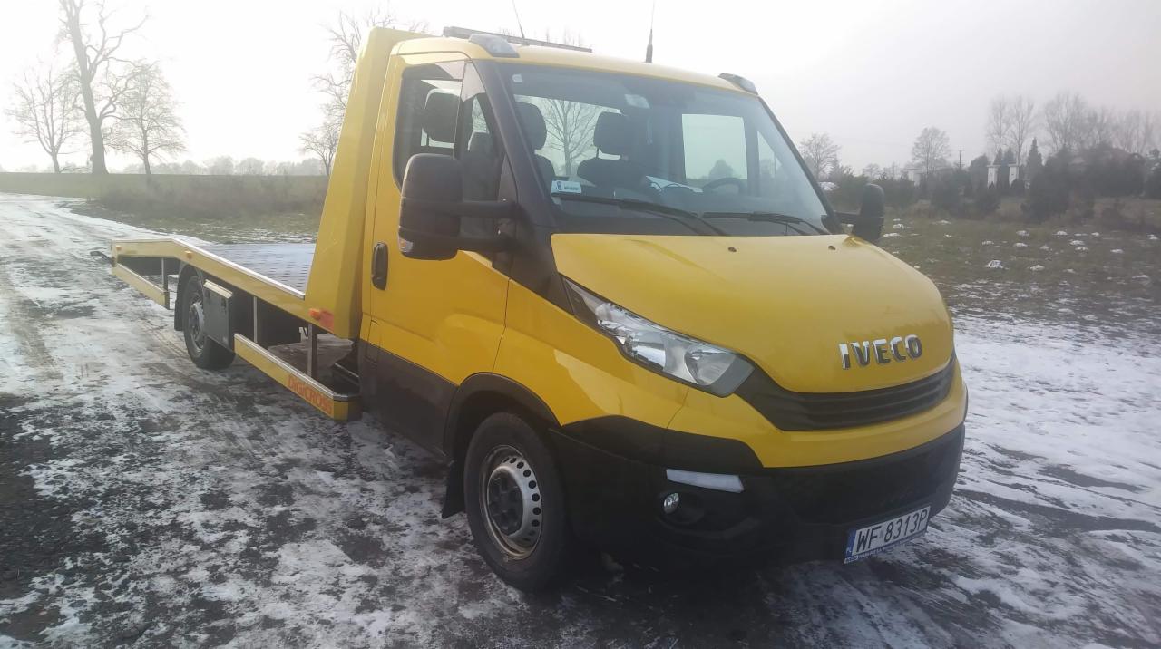 Żółta laweta marki Iveco na poboczu drogi pokrytej śniegiem, widok z przodu na lekko pochmurne tło.