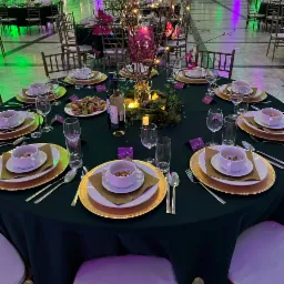 Uroczysty stół bankietowy z ciemnozielonym obrusem, złotymi podkładkami, białą zastawą i dekoracją ze świecących gałązek, przygotowany dla gości z imiennymi wizytówkami.