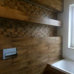 Wykończona ściana w łazience z płytek imitujących drewno, z widocznym przyciskiem spłuczki i fragmentem wanny, uzupełniona mozaiką i półkami.