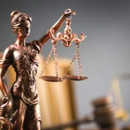 Brązowa statuetka Temidy, bogini sprawiedliwości z zasłoniętymi oczami, trzymająca wagę na tle rozmytych książek i młotka sędziowskiego.